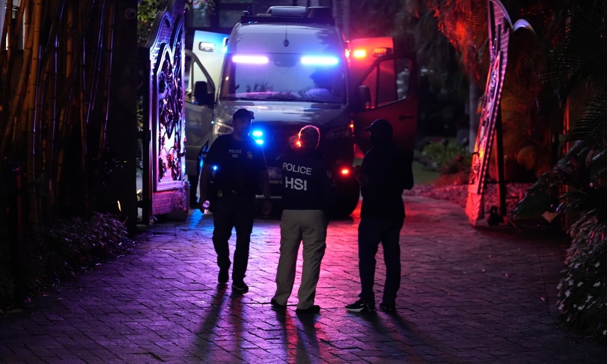 Three police officers stand in a dark alley, lit by the multi-coloured lights coming from a vehicle -- either an ambulance or a large police vehicle -- parked beyond them. 
