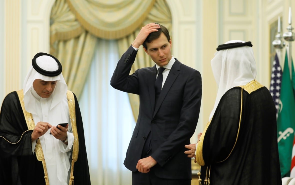 A man in a suit touches his head, flanked by two men in traditional Arabic robes. 