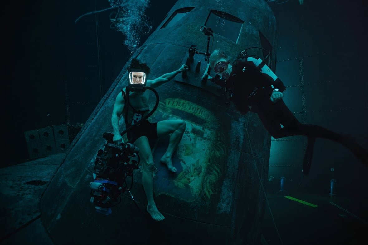 A man wears a diving mask while holding onto the outside of a submarine underwater. He is shirtless and wearing only a bathing suit. Beside him is a diver in full SCUBA gear. 