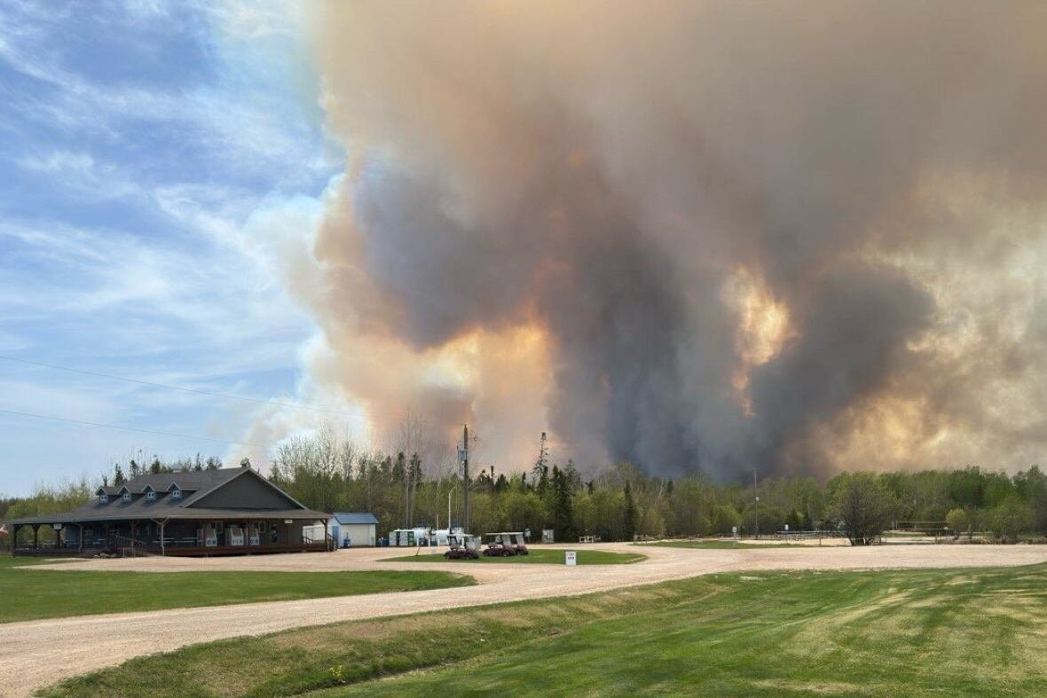 Smoke and flames in the background rise above a forest. A gravel road and store are in the foreground.