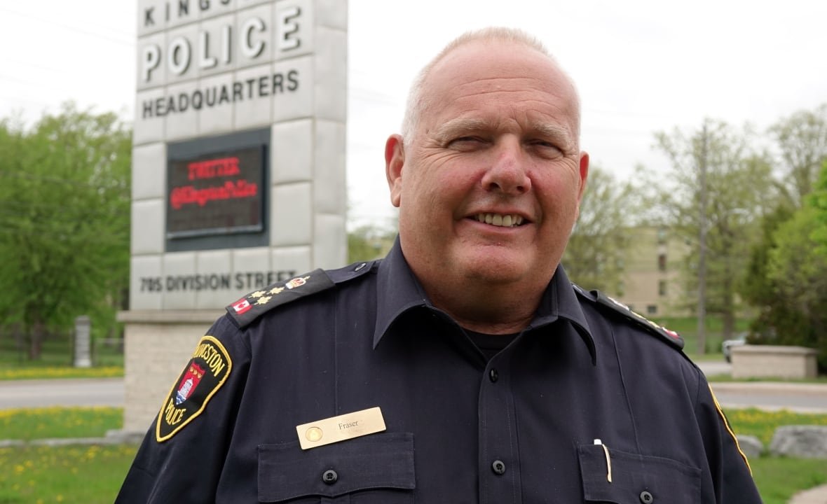 An older man in a police uniform stands in front of a metal sign that says "Kingston Police Headquarters" in large letters.