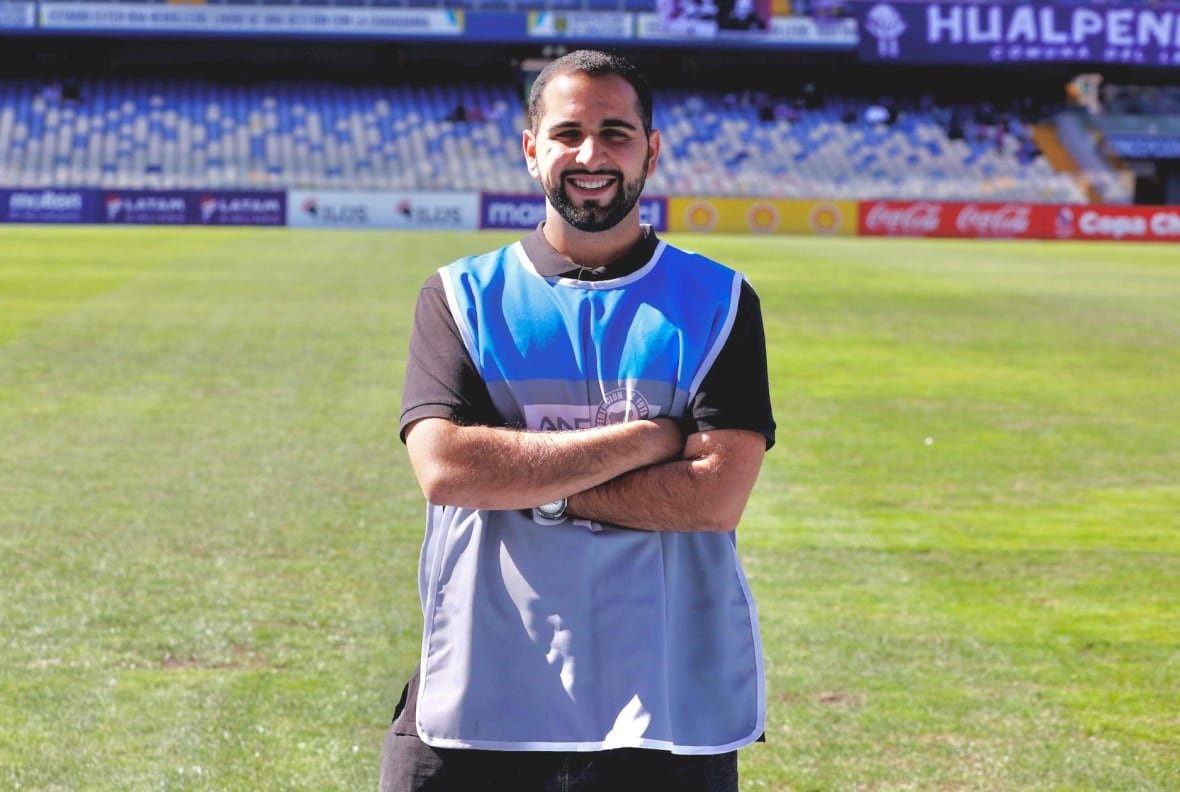 A man with a beard and in a blue and grey press vest stands cross-armed and smiles at the camera.