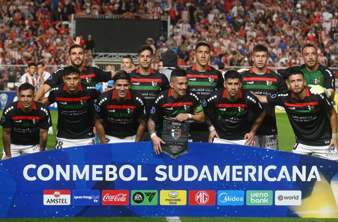 Eleven soccer players, most in black jerseys and white shorts, stand in front of a board reading "CONMEBOL Sudamericana" and some sponsors. 
