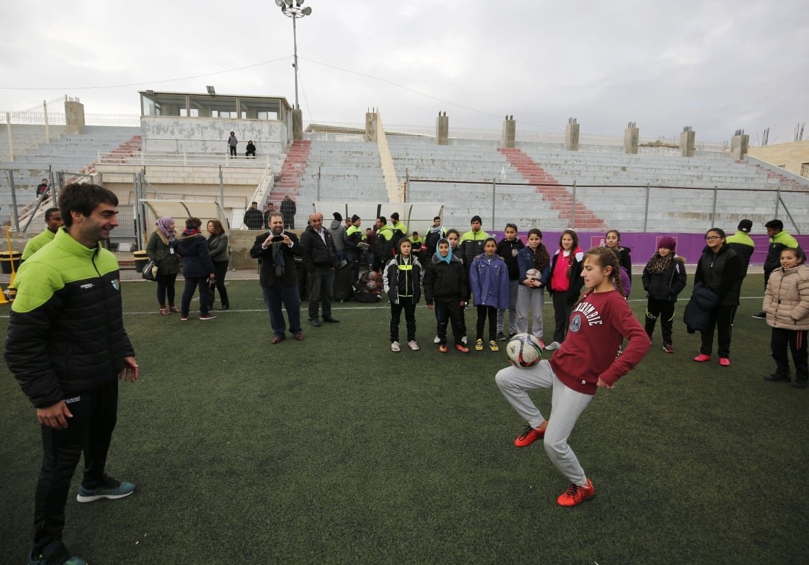 A girl in a maroon sweater and grey pants juggles a soccer ball. She's surrounded by other children and members of the Palestino soccer team.