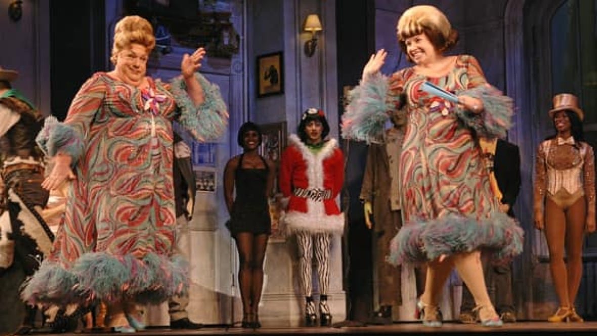 A man wearing a very loud print dress and large wig performs on stage next to a woman in a similar outfit. 