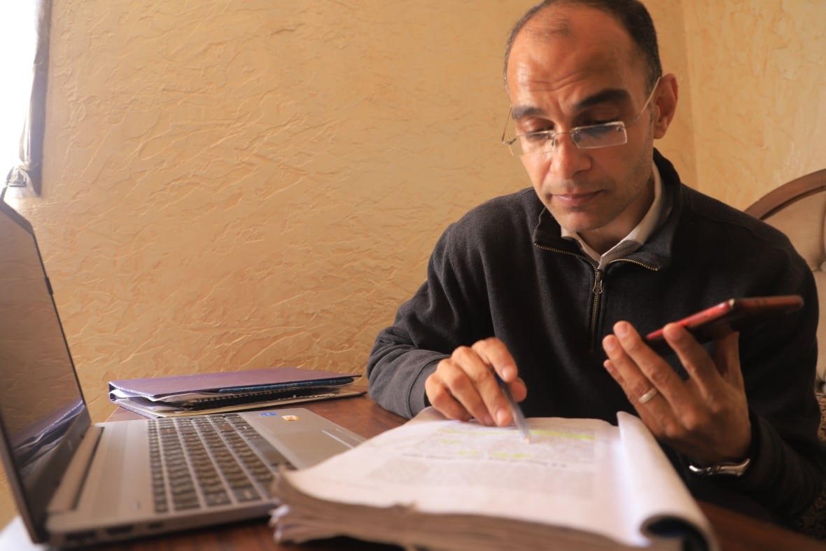 a balding man with glasses sits at a desk with a cellphone, notebook and laptop 