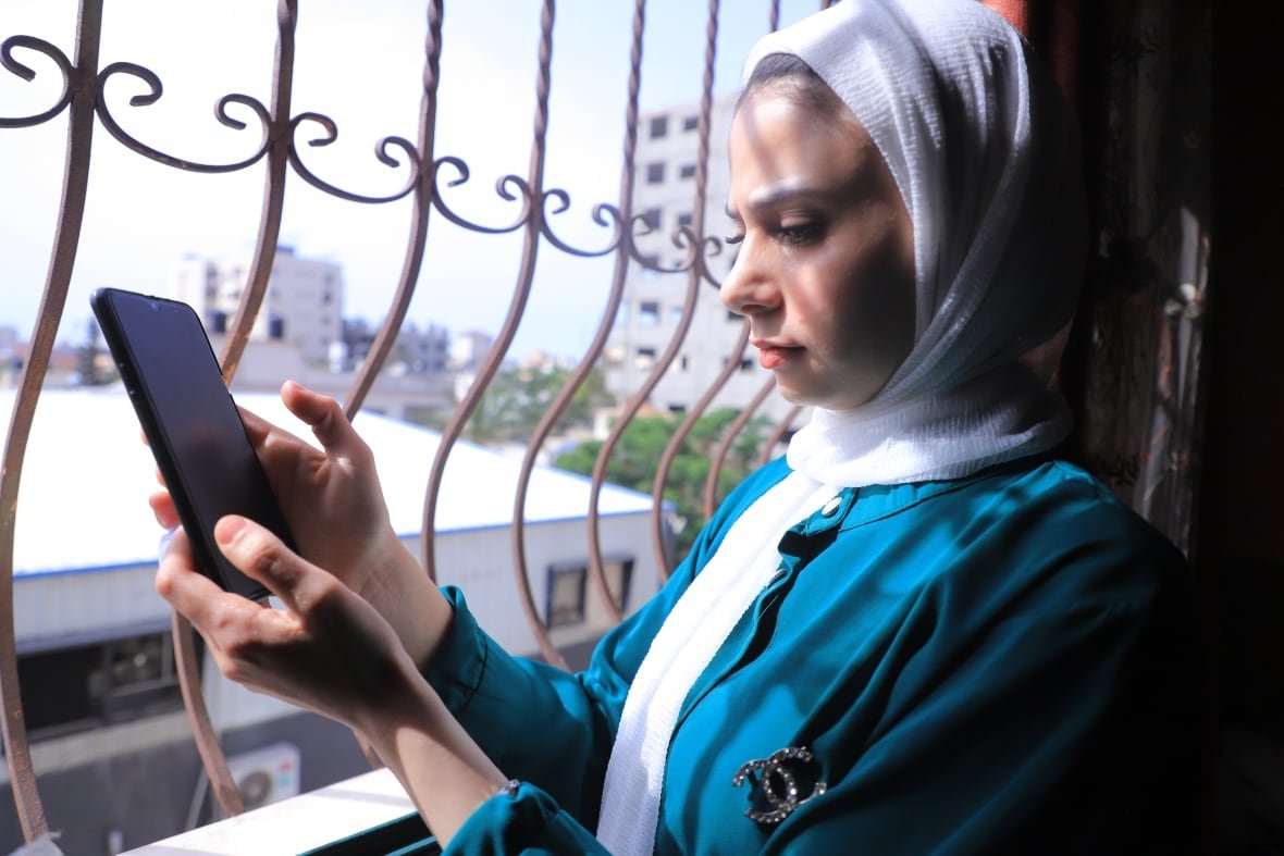 a girl in a white hijab and a blue shirt holds a cellphone as she stands next to a window with a cast-iron railing. 