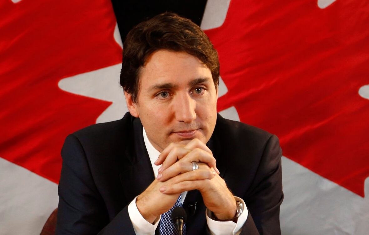 A man, seated in front of some Canadian flags, leans forward, resting his chin on his folded hands.
