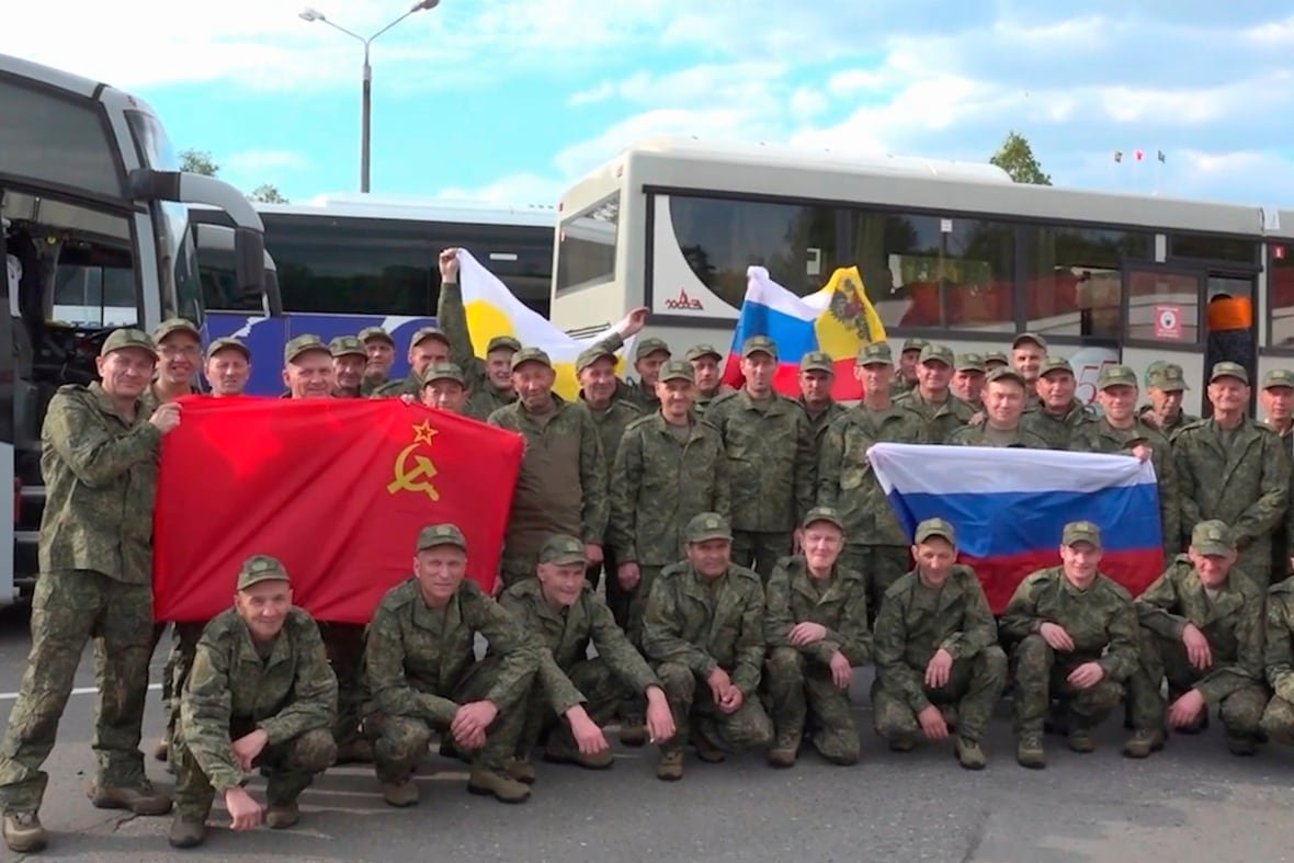 Dozens of military members in camouflage style uniform pose with flags. Some are crouching, some standing.