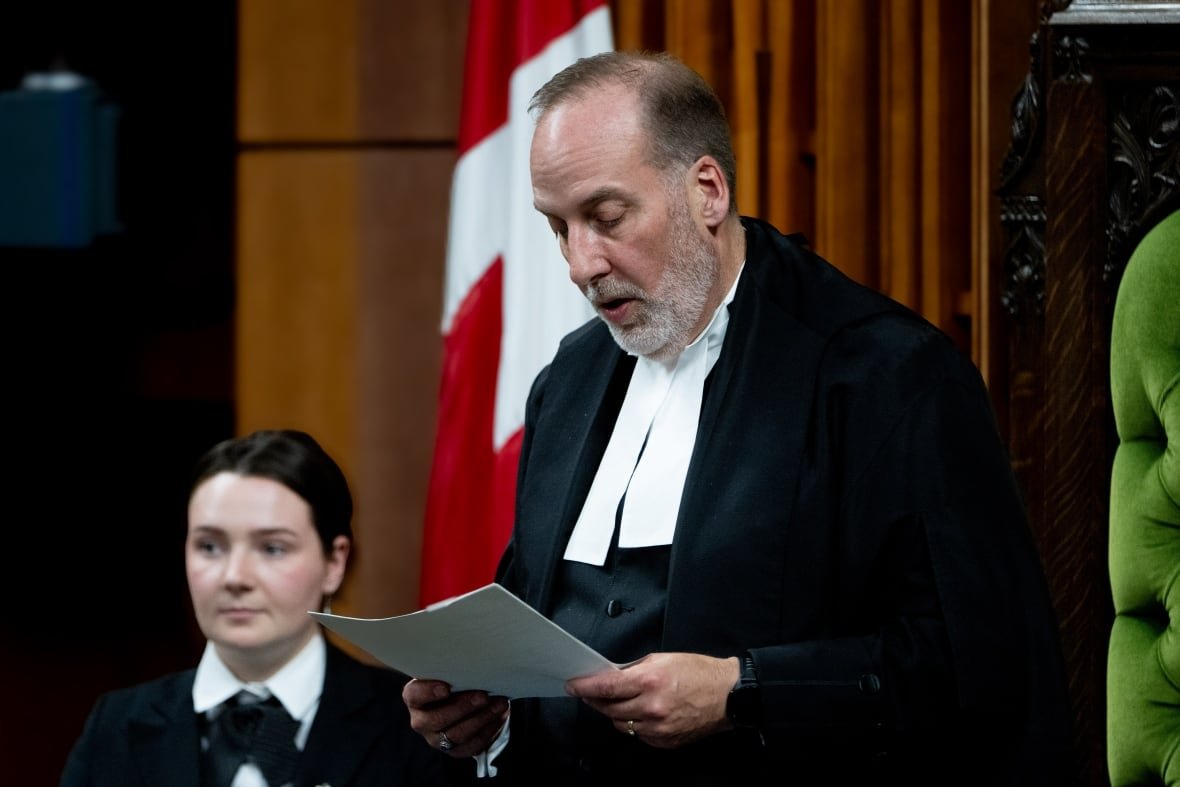 A man in a formal House of Commons gown reads off of a piece of paper.