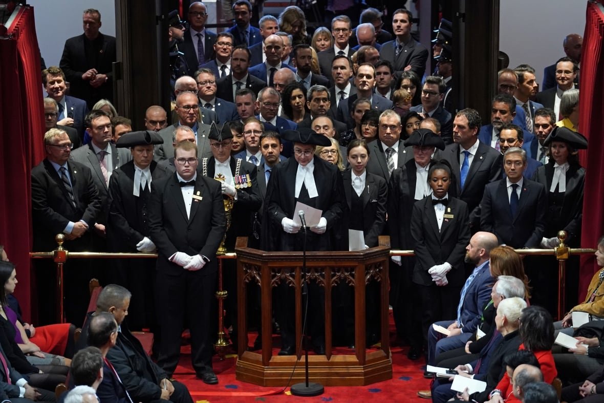 A crowd of people stand in front of a brass bar in Canada's Senate.