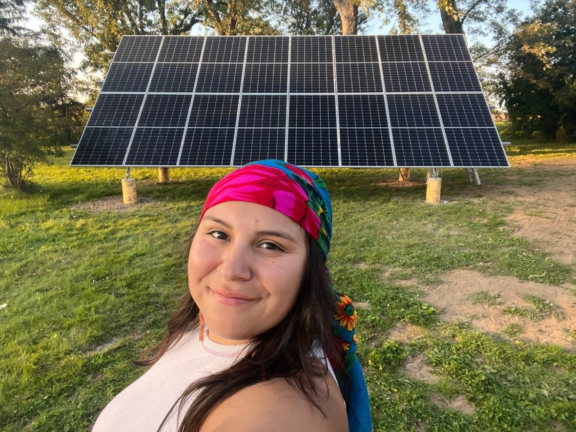 A young Indigenous woman in a bright pink and blue headscarf poses with a large block of solar panels standing behind her. She smiles.