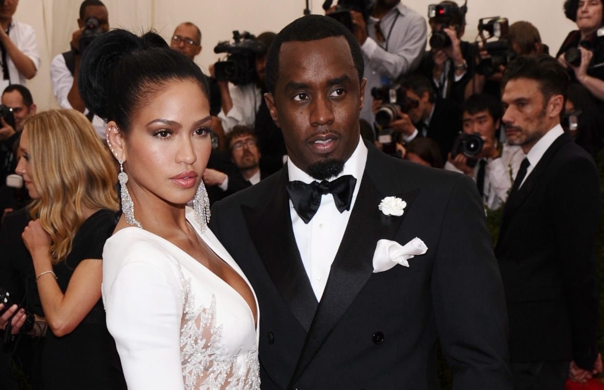 A man and a woman pose together on a red carpet. They are shown from the waist up and are posing turned in towards each other, looking out. The man is in a black suit an the woman is in a white, long-sleeved dress.