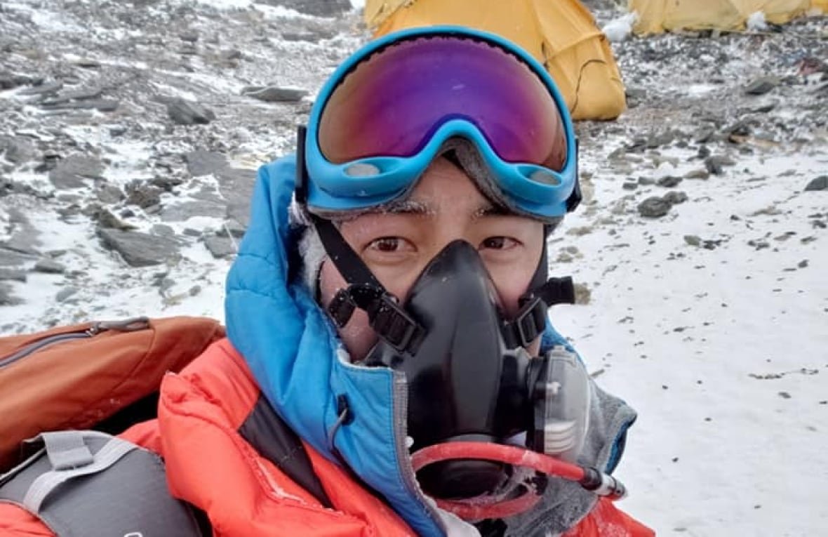 A man wearing ski goggles, an orange parka and a black respirator takes a selfie on the side of a mountain. 