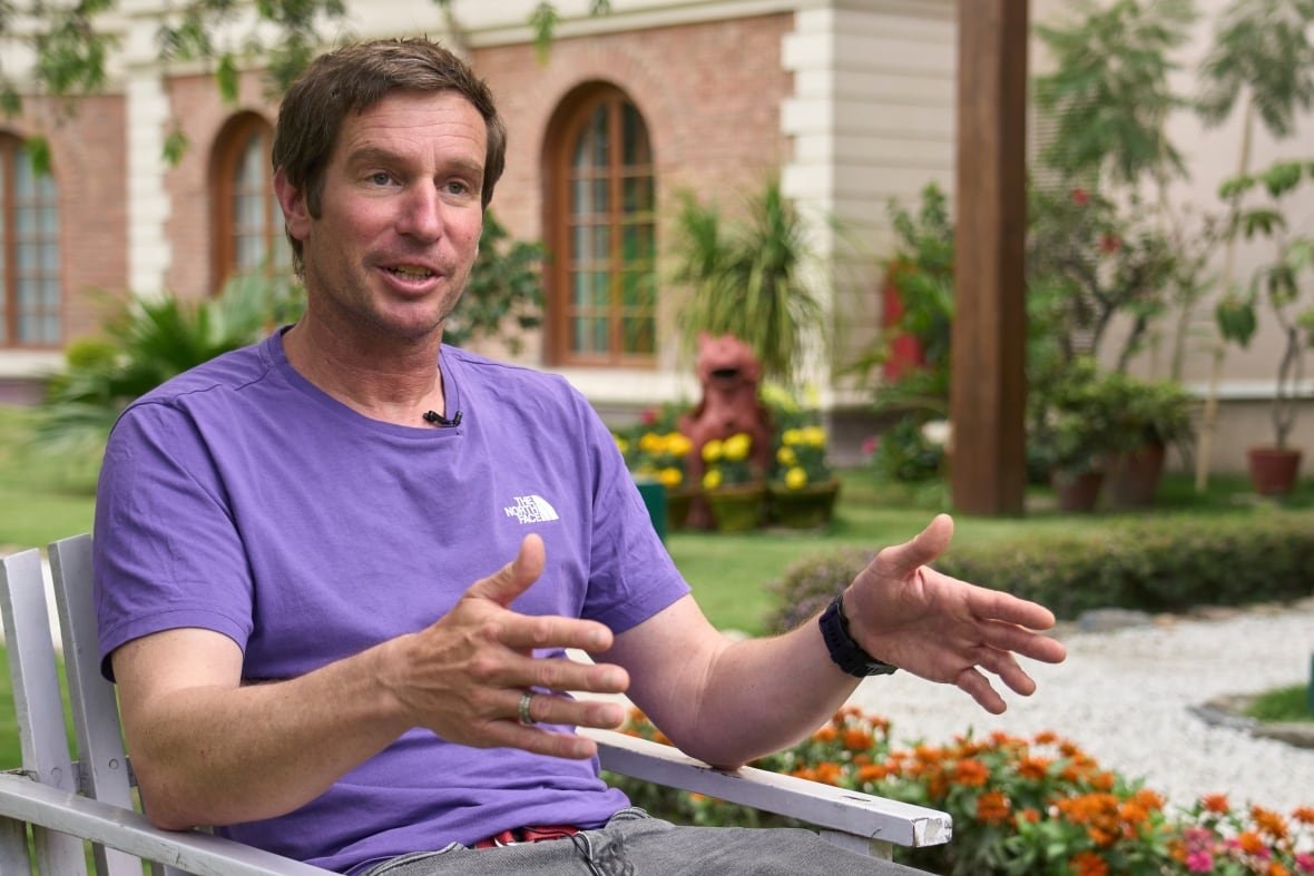 A man in a purple t-shirt speaks while sitting on a wooden chair in a courtyard garden. 