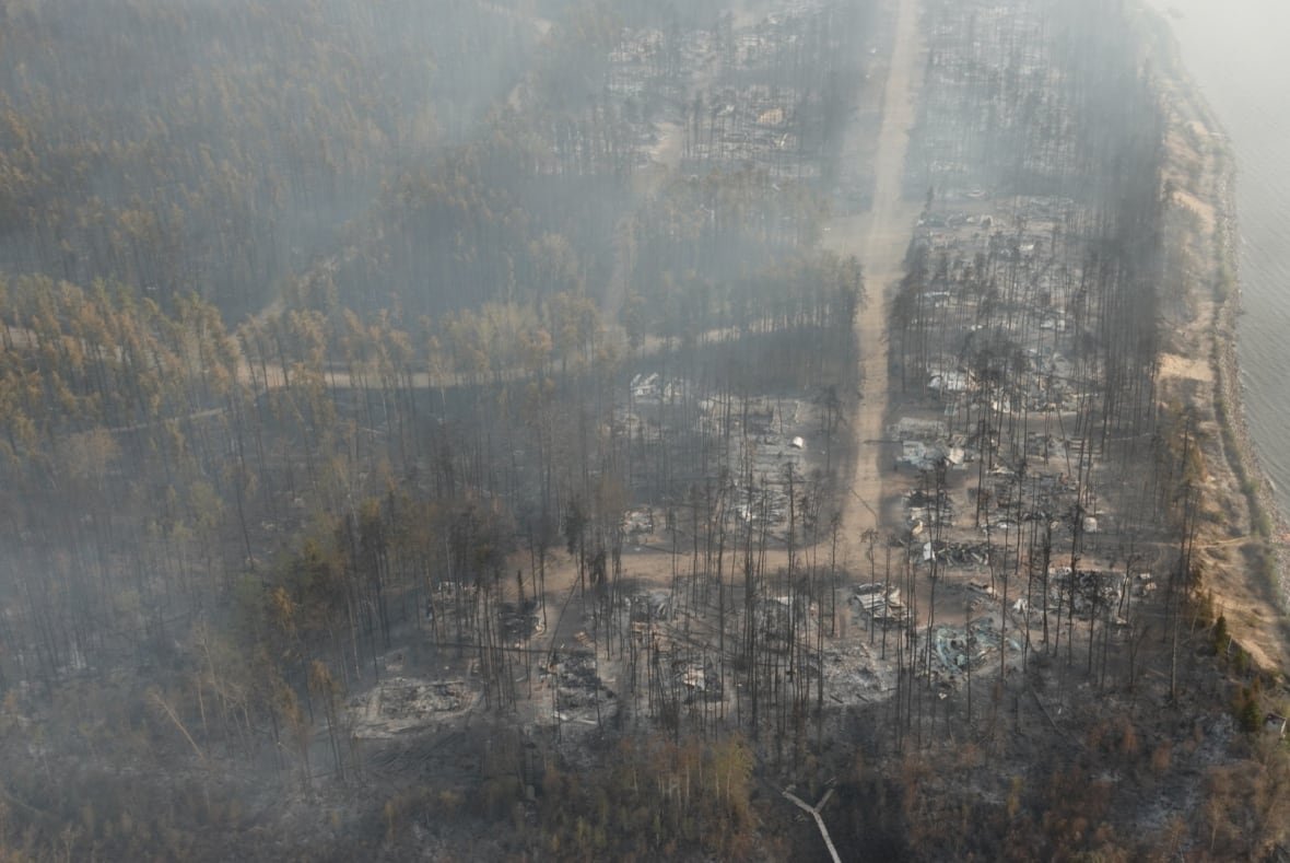 Burned trees stand near a lake. Charred remains of cabins can be seen