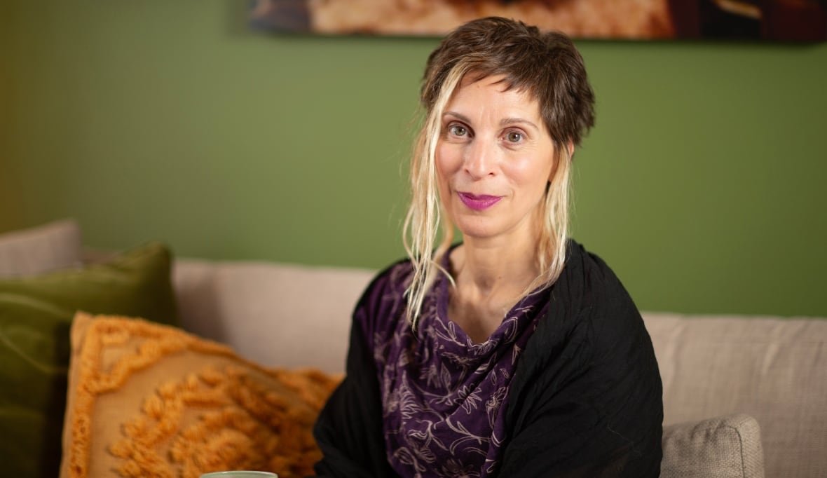 A woman with short dark hair highlighted by long blond streaks sits on a beige couch in front of a green wall with throw pillows to her side. 