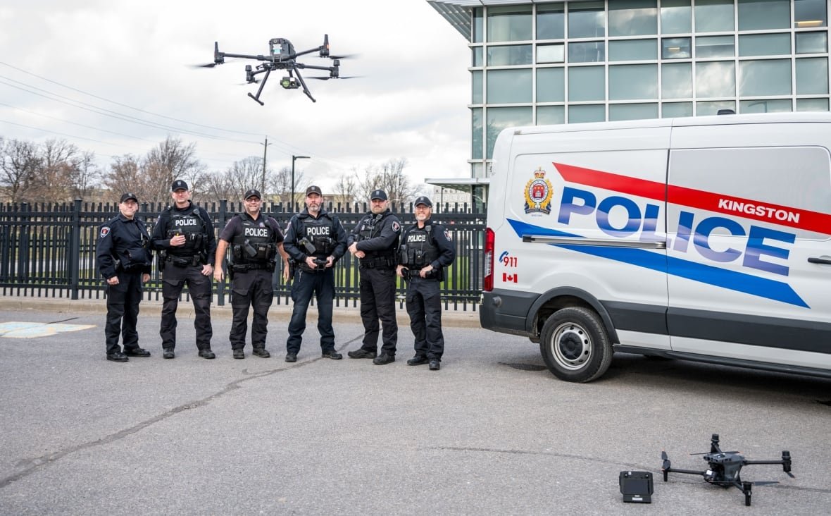 Six police officers in full uniform stand next to a white van with the Kingston Police logo on it. One of the officers is flying a drone, which appears above them.