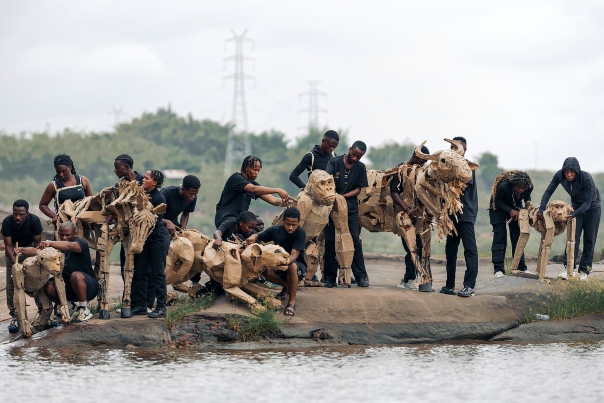 People dressed in black at a pond with cardboard animal puppets, including a gorilla and a gnu.