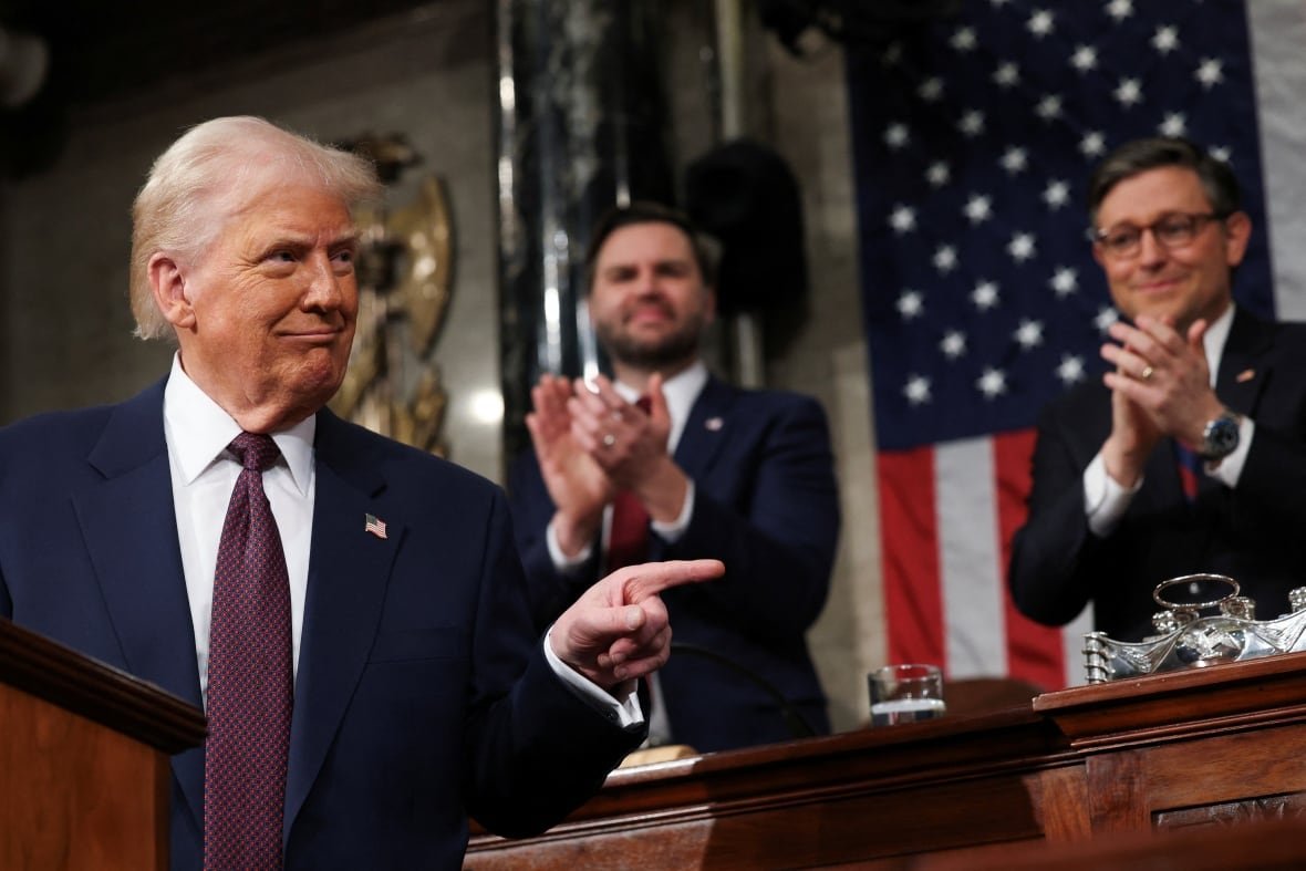 A man in a suit points a finger as two other men applaud behind him in front of an American flag.