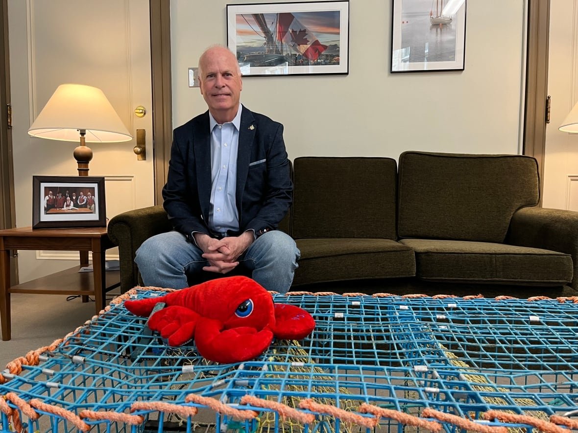 A bald man sits on a green couch. In front of him is a stuffed lobster on top of a trap.