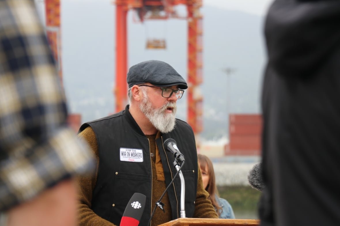 A man with a black hat, glasses and grey beard speaks into a microphone on a beach along Vancouver's downtown waterfront.