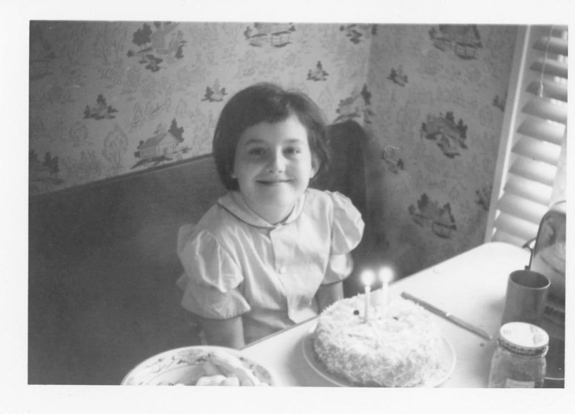 A black and white image of a girl, smiling at the camera, with a bob, an old-school dress. She is sitting at a kitchen booth. There is a cake with candles in front of her.