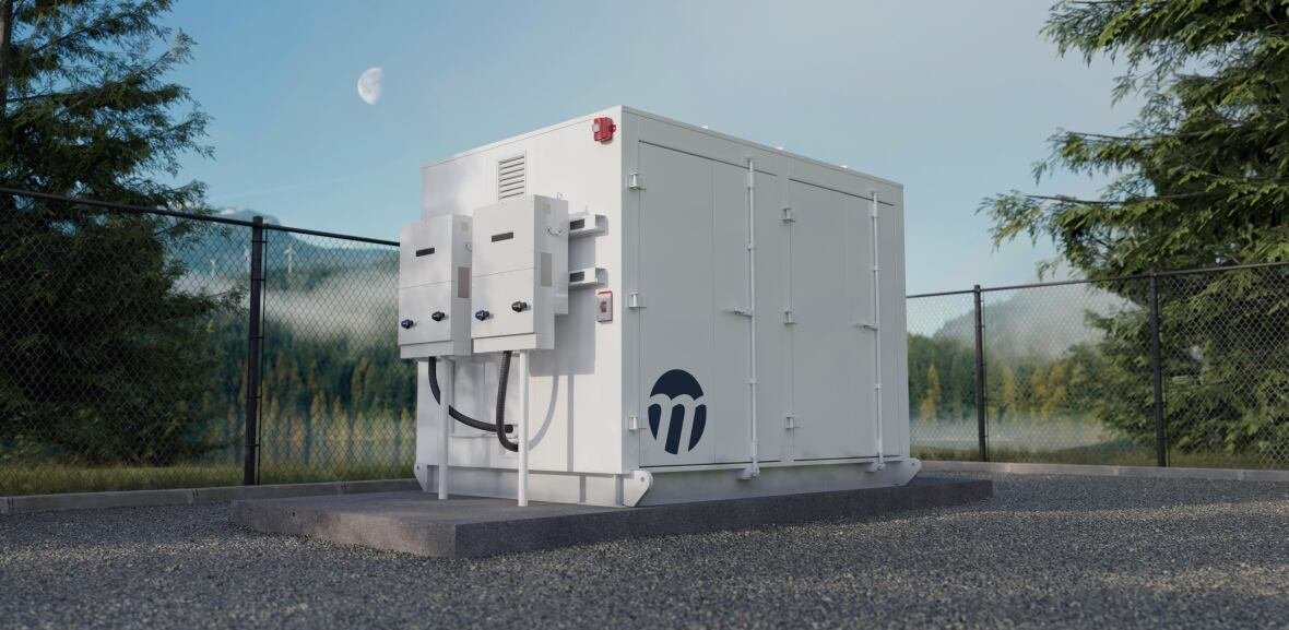 A white box, around the size of a small car, is sitting in a gravel lot with a wire fence in the background and trees beyond. 