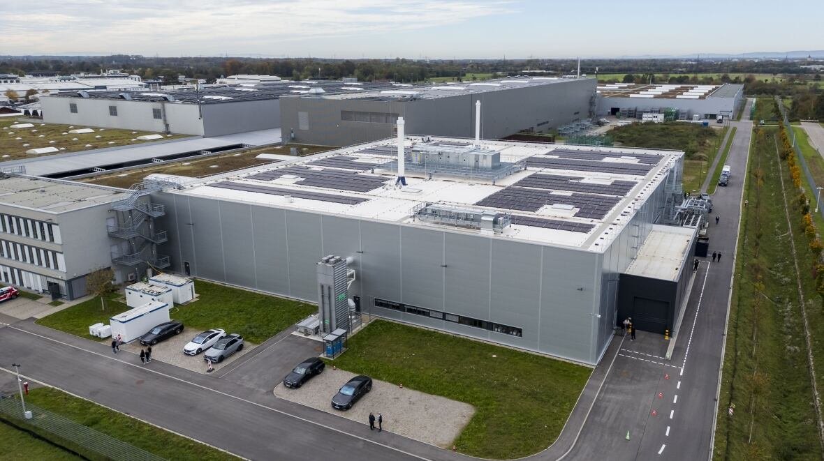 An aerial shot shows a large factory building near a road. 