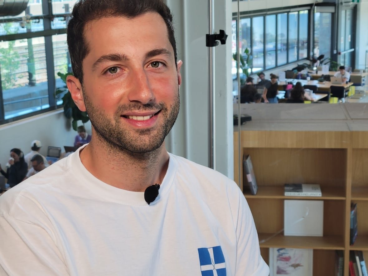 A person from the chest up, with office workers and a bookshelf in the background. 
