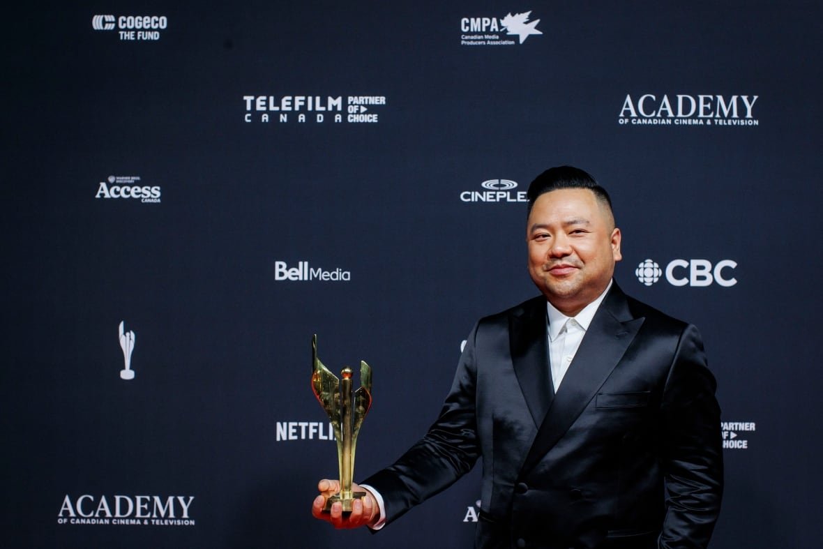 A man wearing a black suit holds out a golden trophy.