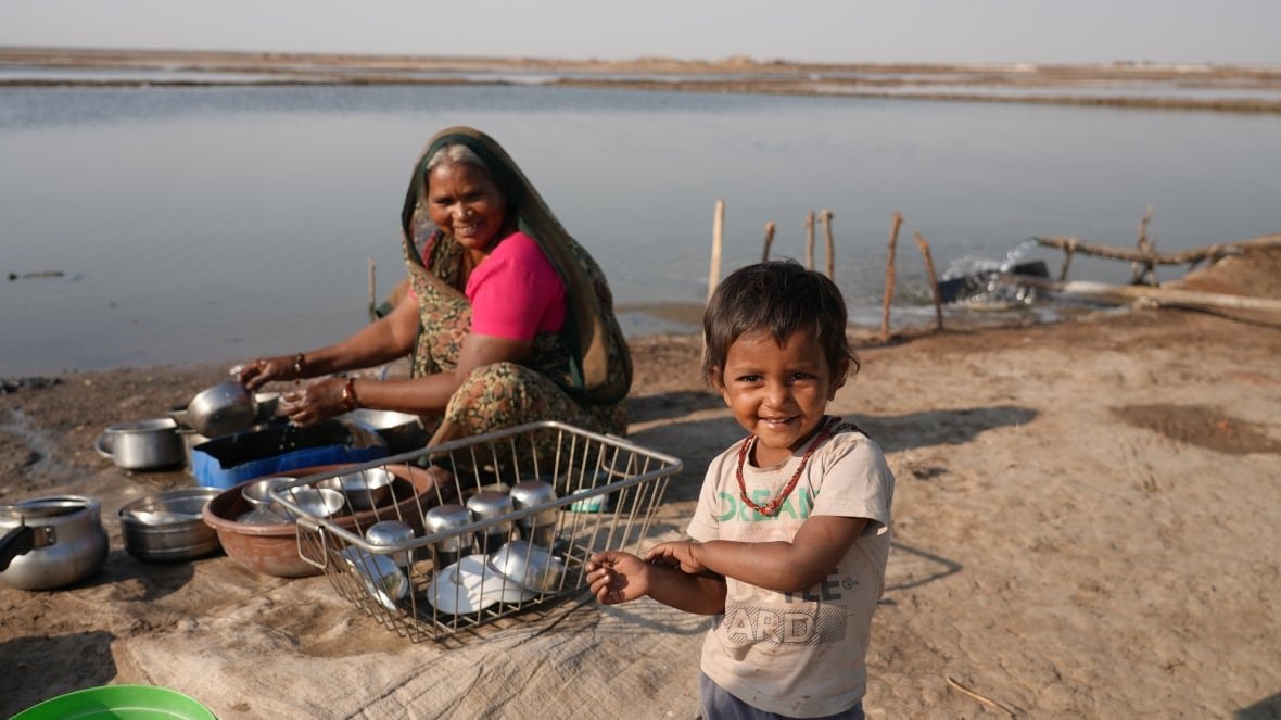 A person seated on a beach washes pots with a child in the foreground.