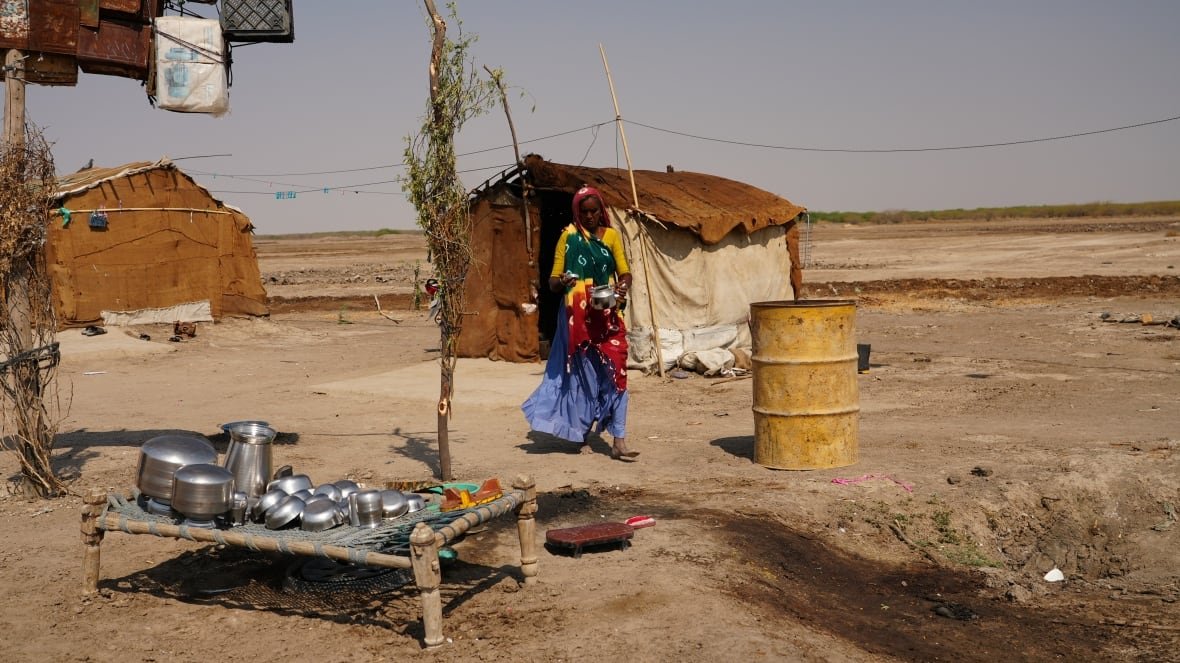 A person holding a pot walks toward a barrel, with tents in the background and a low table with pots in the foreground.