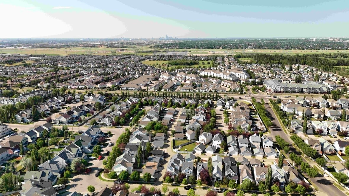 A suburban neighbourhood sits under a cloudless sky. Vibrant green trees line the sidewalks. The skyline of downtown Edmonton is visible in the background.