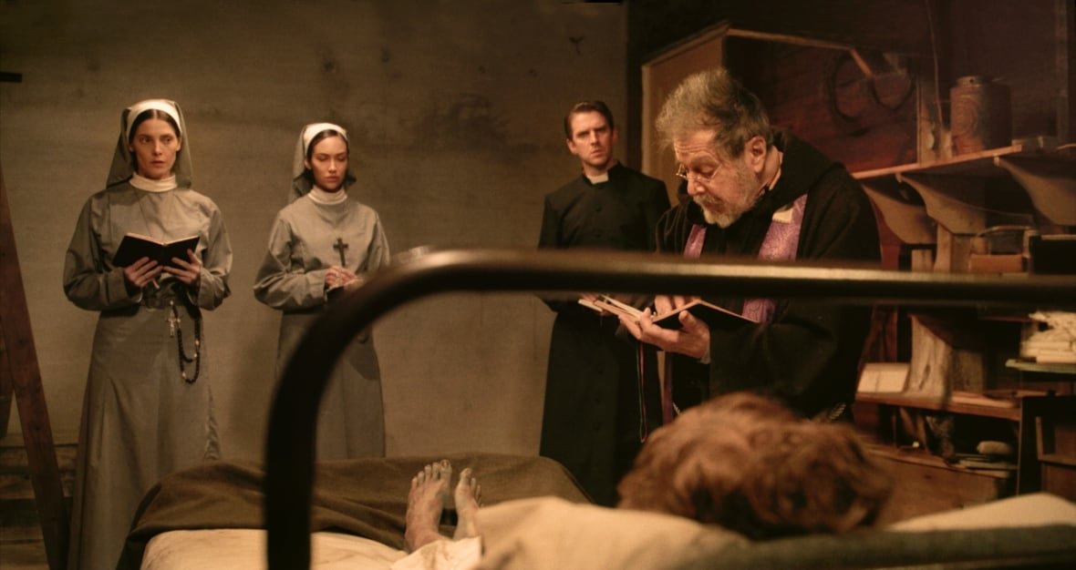 A priest reading from a book stands in front of a woman in a bed and beside two nuns and another priest.. Only the woman's feet are visible, which are bare and dirty.