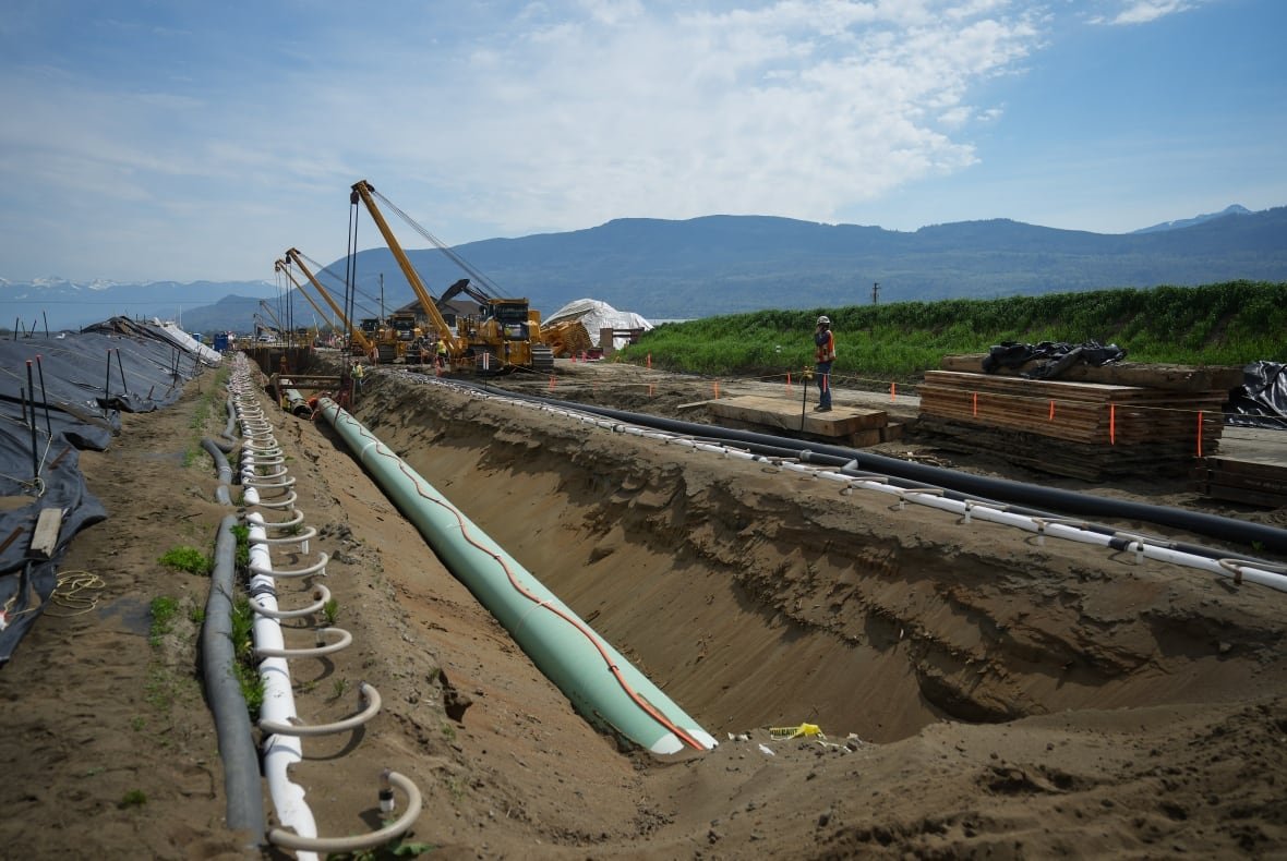 a construction site in a remote natural area shows a worker standing near a large pipeline with a mountain range in the background.