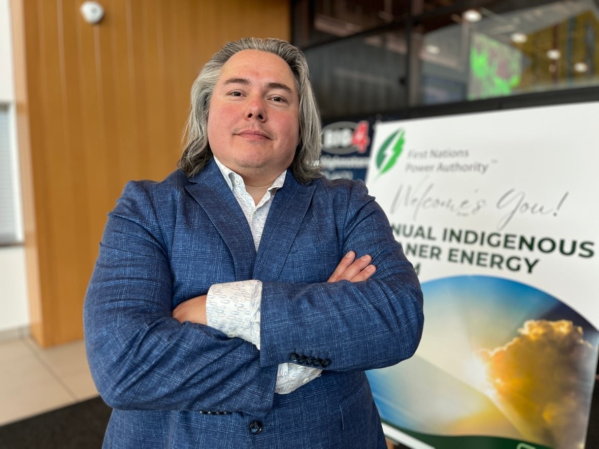 A man with long hair, a blue blazer and a dress shirt is pictured with his arms crossed in front of a conference sign.