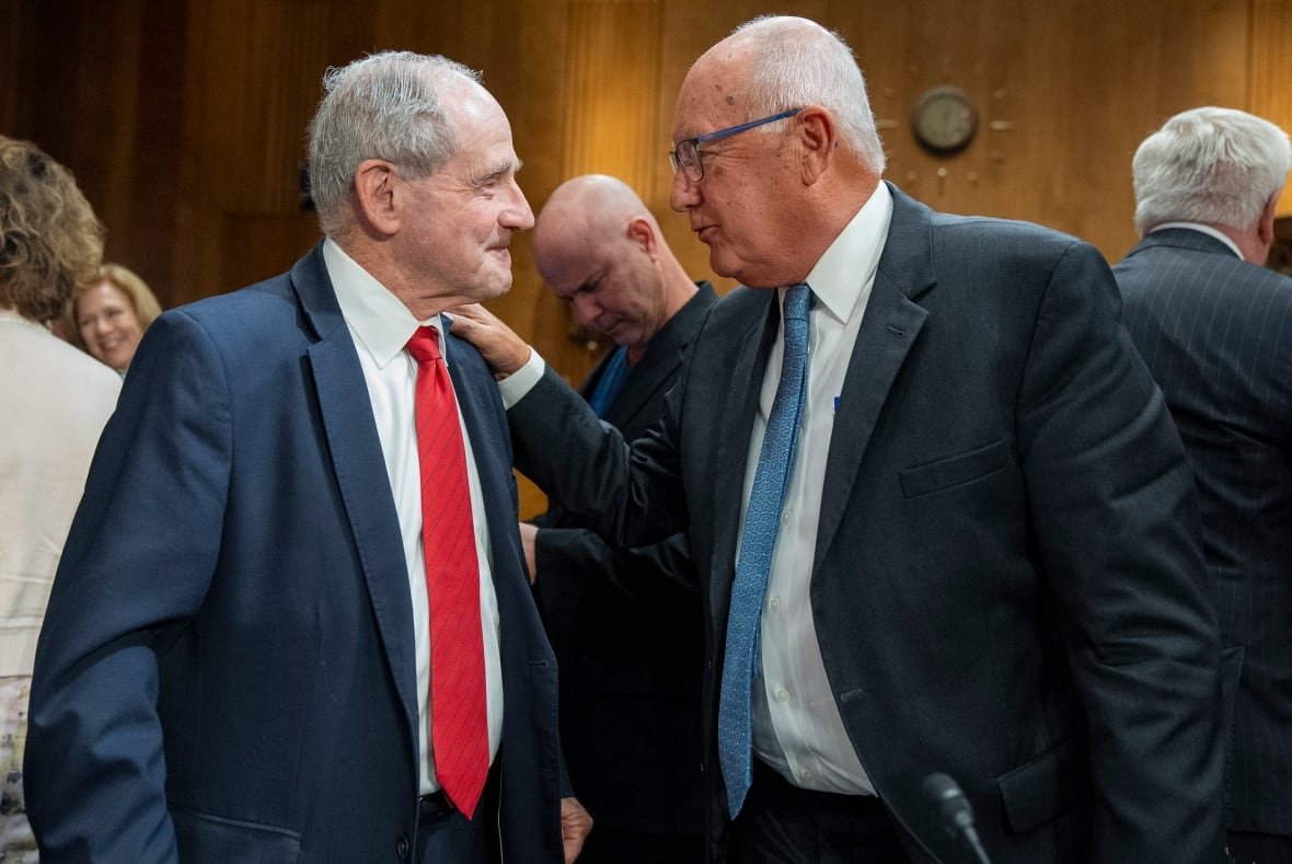 Two men in suits face each other, one with his hand on the other's shoulder.