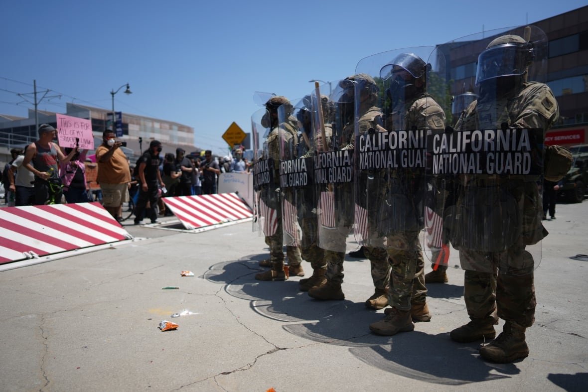 Five uniformed members of the U.S. National Guard stand with their shields up as protesters stand behind a barrier to the left. 