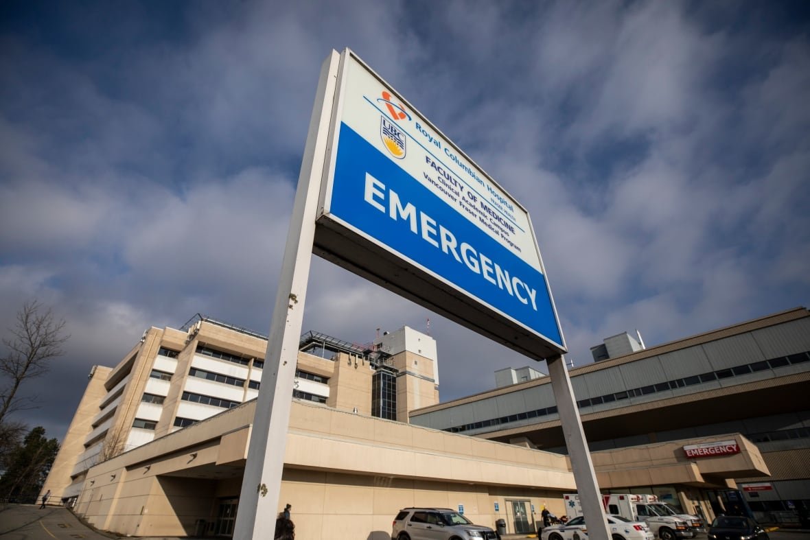 A blue sign with "EMERGENCY" in white capital letters is pictured outside a beige hospital on a stormy day.