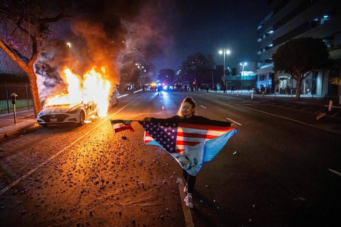 A woman extends an American flag while walking in the middle of a street, with a burning vehicle nearby.