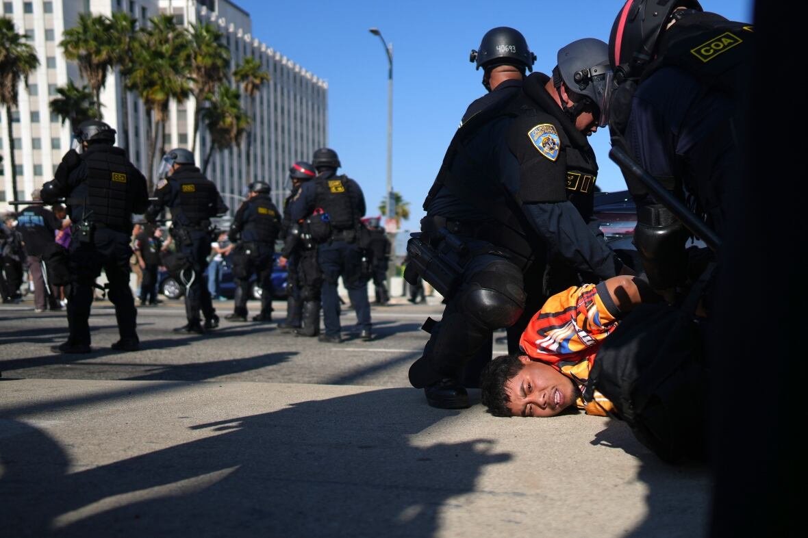 A man in a colourful shirt grits his teeth as his face is pushed into the pavement as police officers in black uniforms kneel next to him and cuff his hands behind his back. 