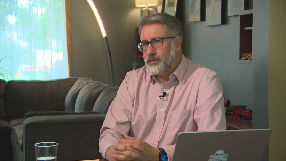A man sitting in a living room wears a pink button-up shirt.