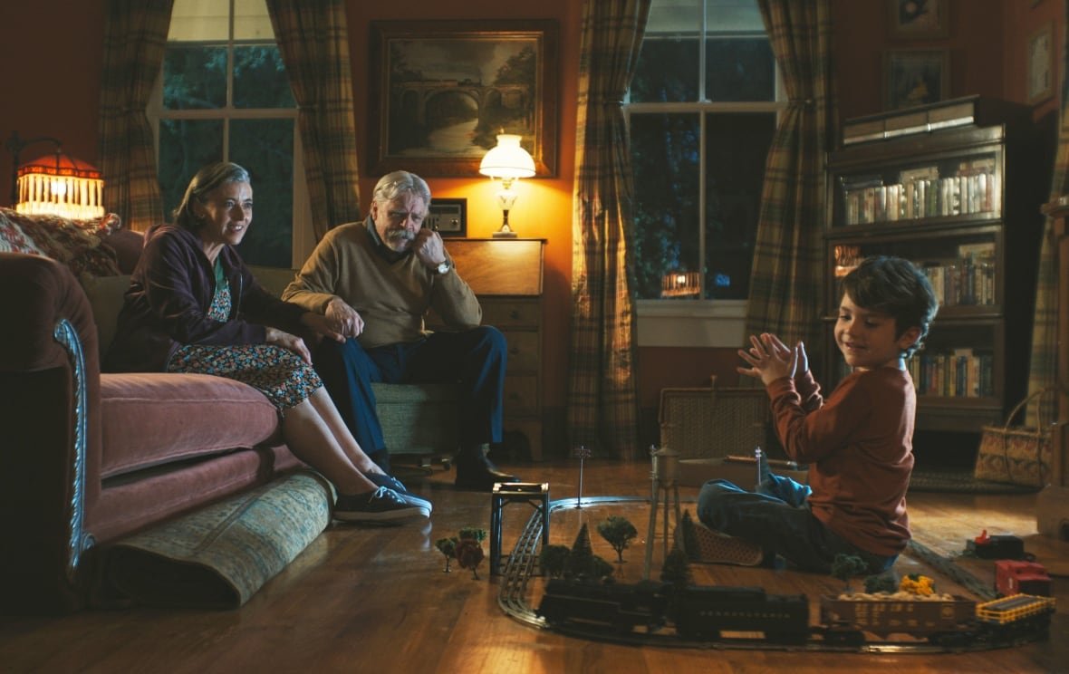 A smiling little boy on the right plays with model trains. Seated to his left are a smiling old man and woman. 