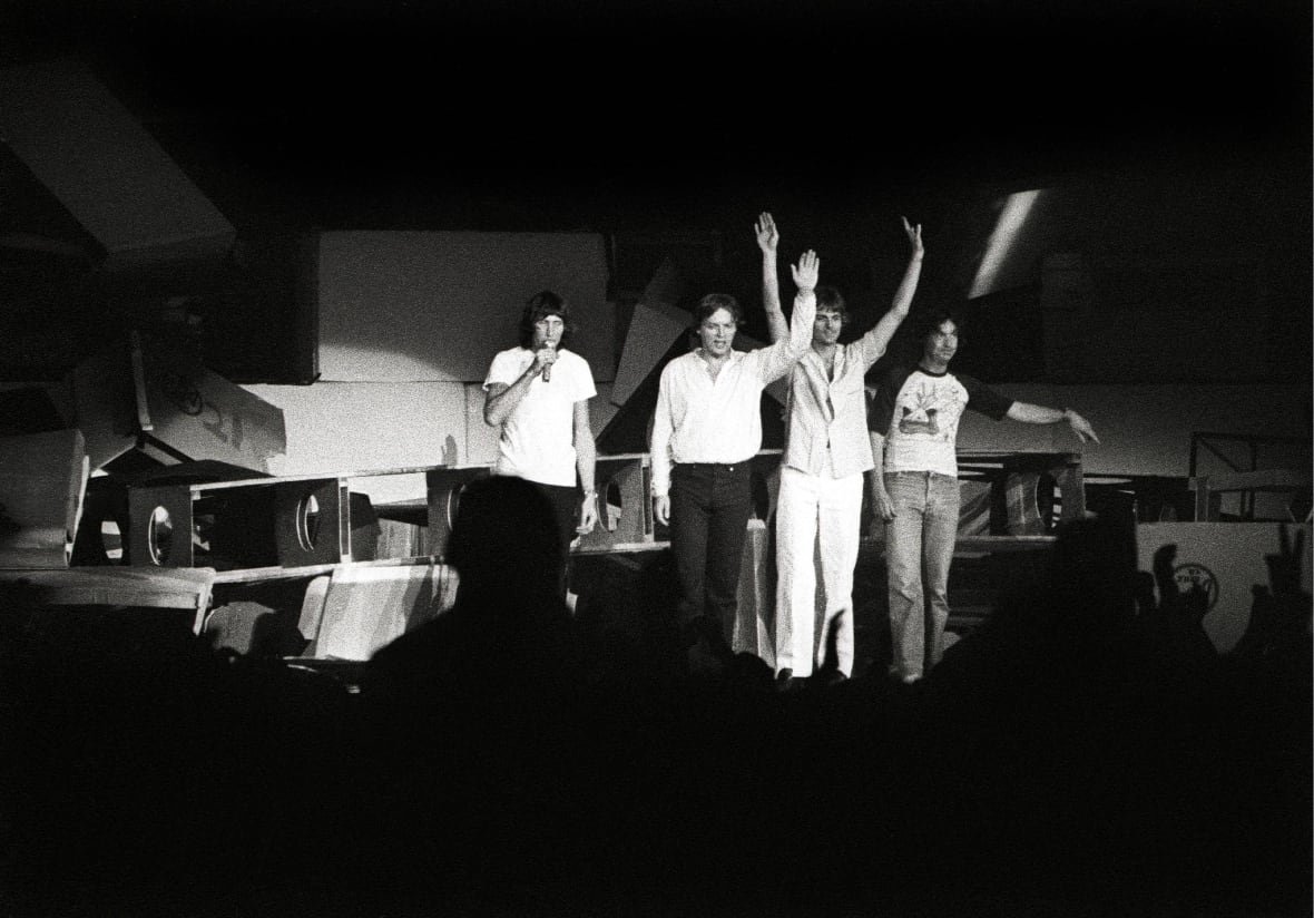 Four men stand on stage and speak to the audience in this black-and-white photo.