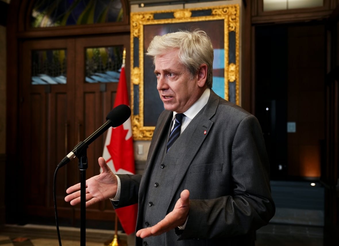 A man in a suit gestures while speaking at a microphone. There is a Canadian flag behind him. 