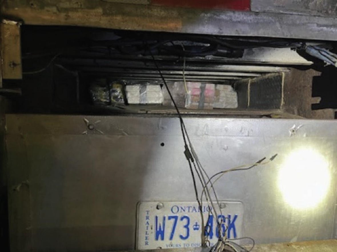 A secret compartment is seen in the back of a grey truck, with packages of what appear to be bricks of drugs