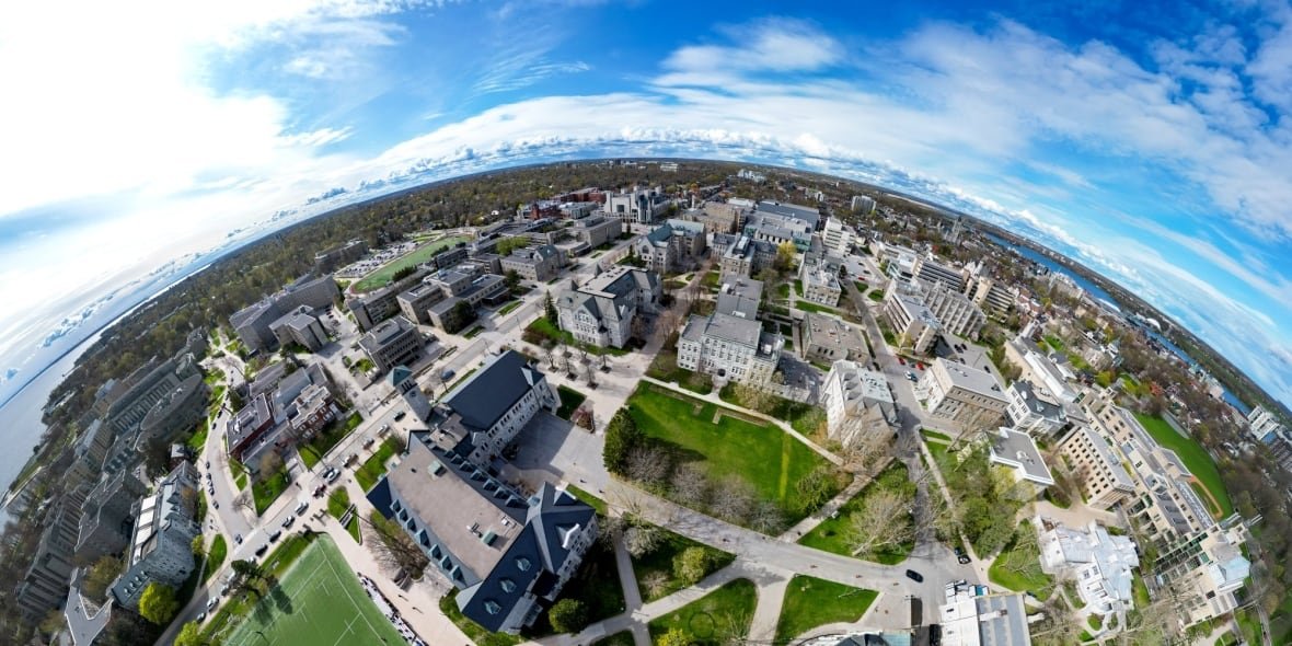 An aerial panorama shows a university campus, with a big blue sky and many grey and brown buildings on green grass. 