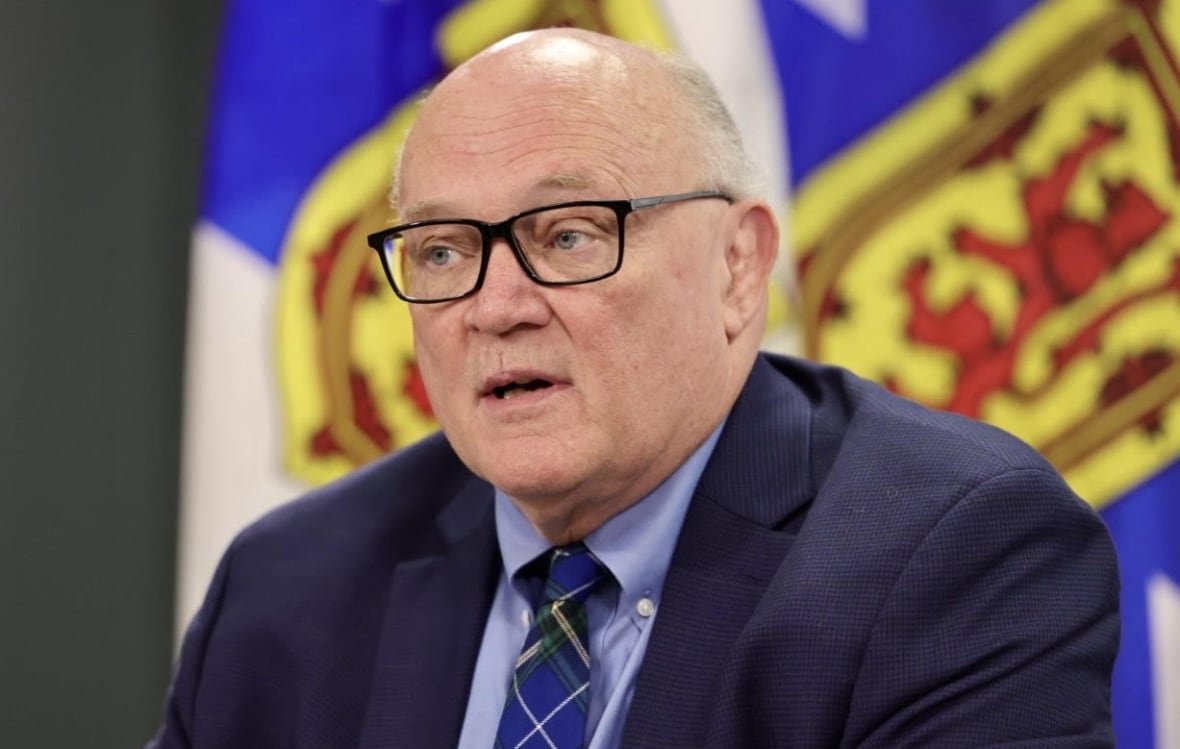A man with glasses and wearing a blue jacket, shirt and tie, speaks in front of Nova Scotia's provincial flags.