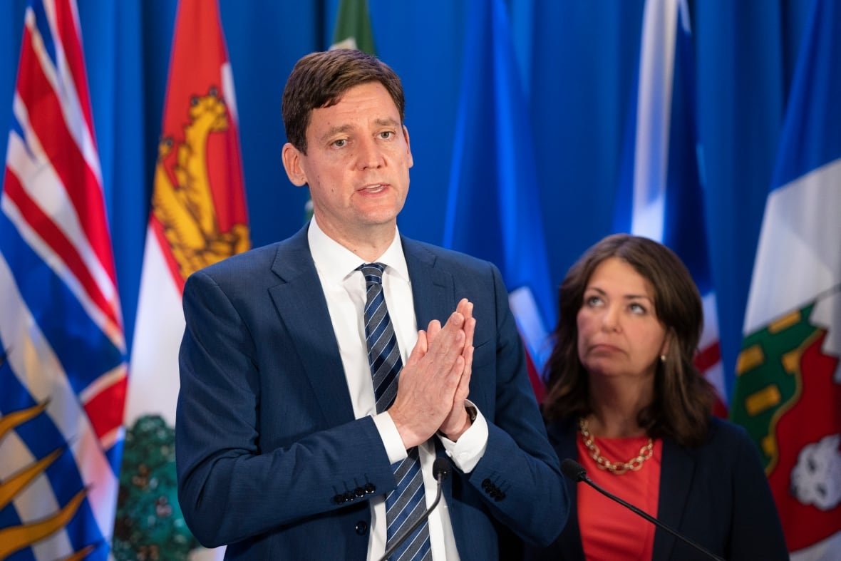 A tall white man folds his hands in front of a series of provincial flags, while a white woman looks up at him.